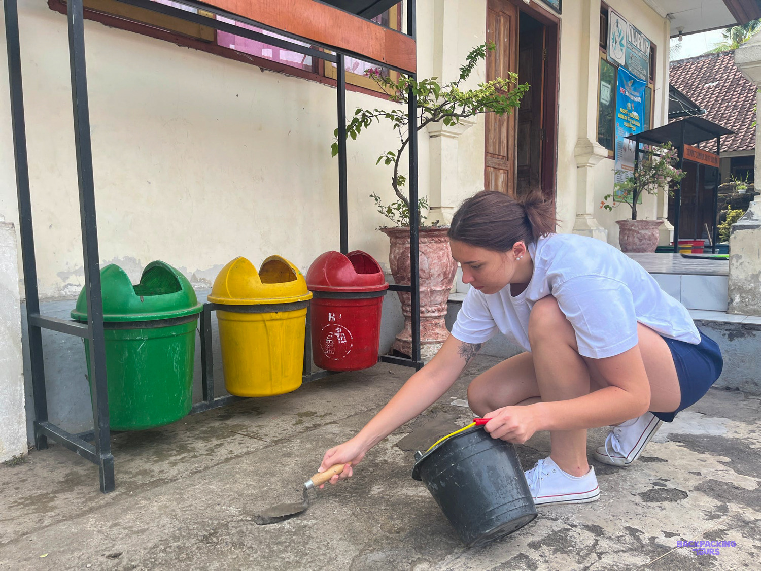 A volunteer kneels to paint the base of a stand of coloured recycling bins at a school.