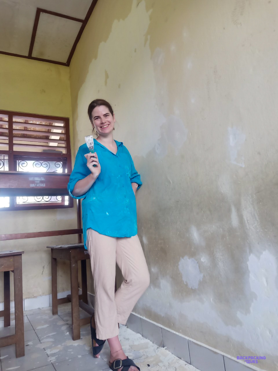 A volunteer stands in a classroom beside a patched and plastered wall ready for painting.