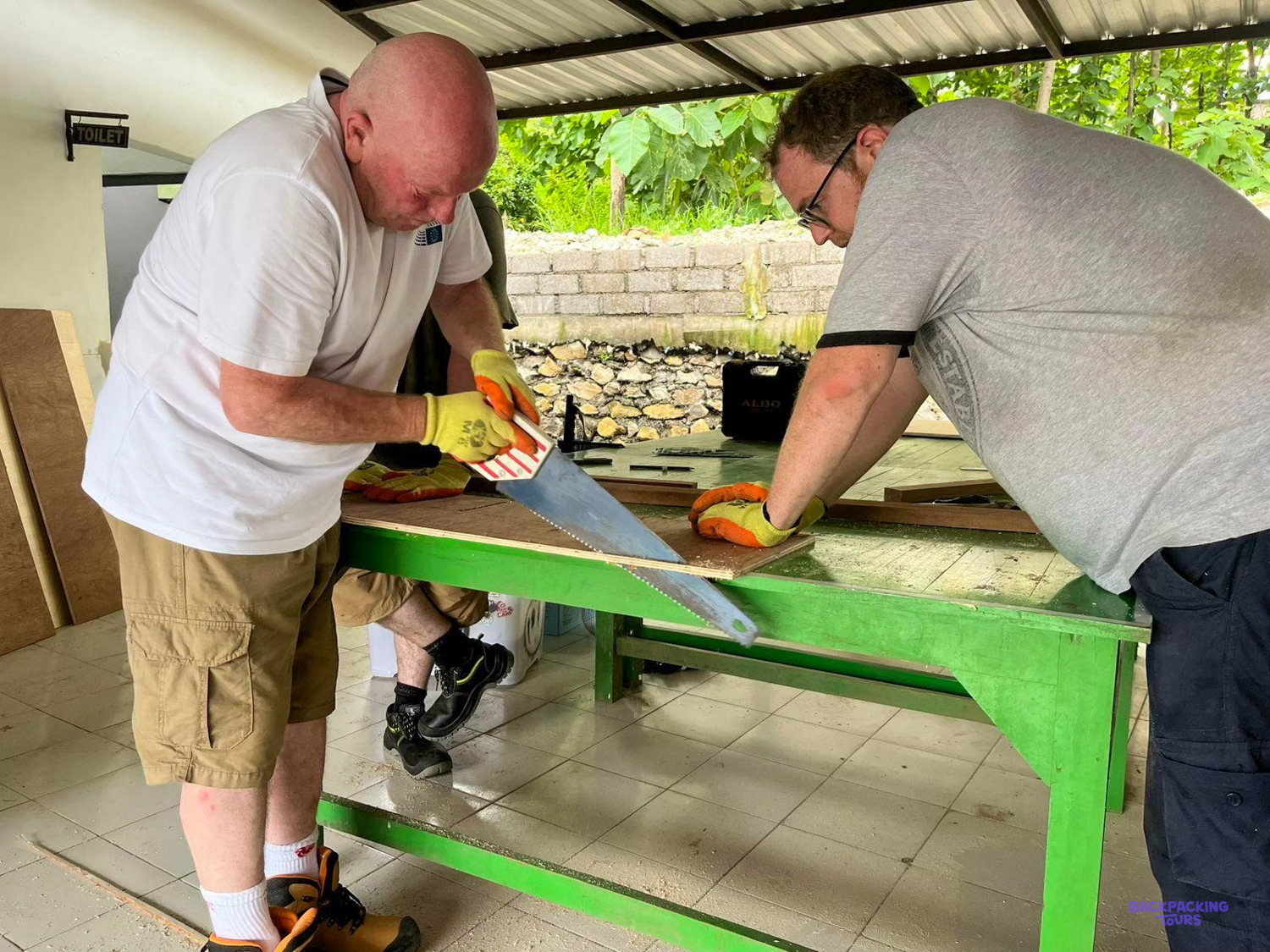 Two volunteers saw and measure a board on a green workbench under a school canopy.