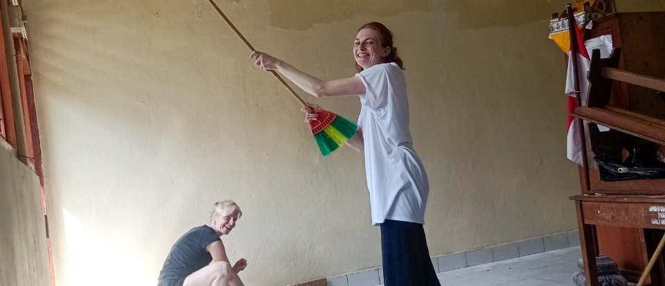 A volunteer paints a classroom wall with a long roller while another works on the floor.