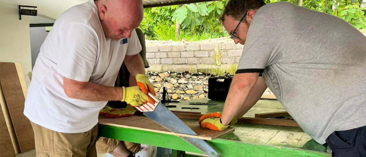 Two volunteers saw and measure a board on a green workbench under a school canopy.