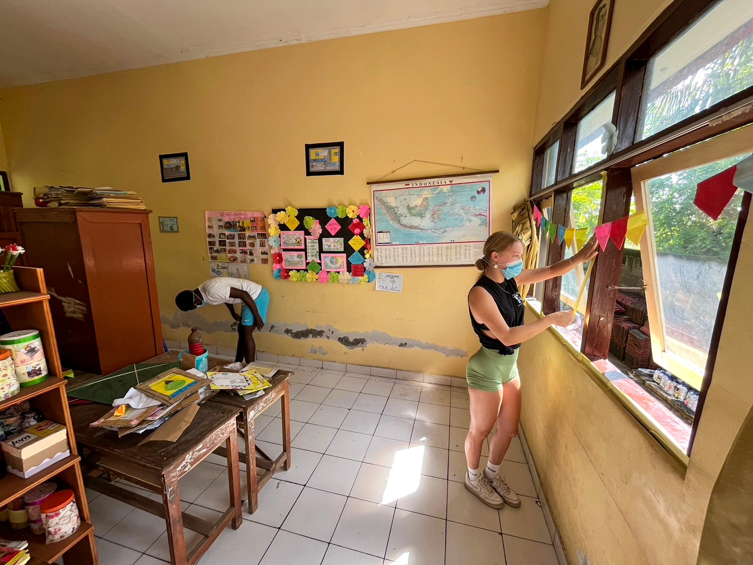 A volunteer tapes up a classroom window frame for painting while another works along the wall.