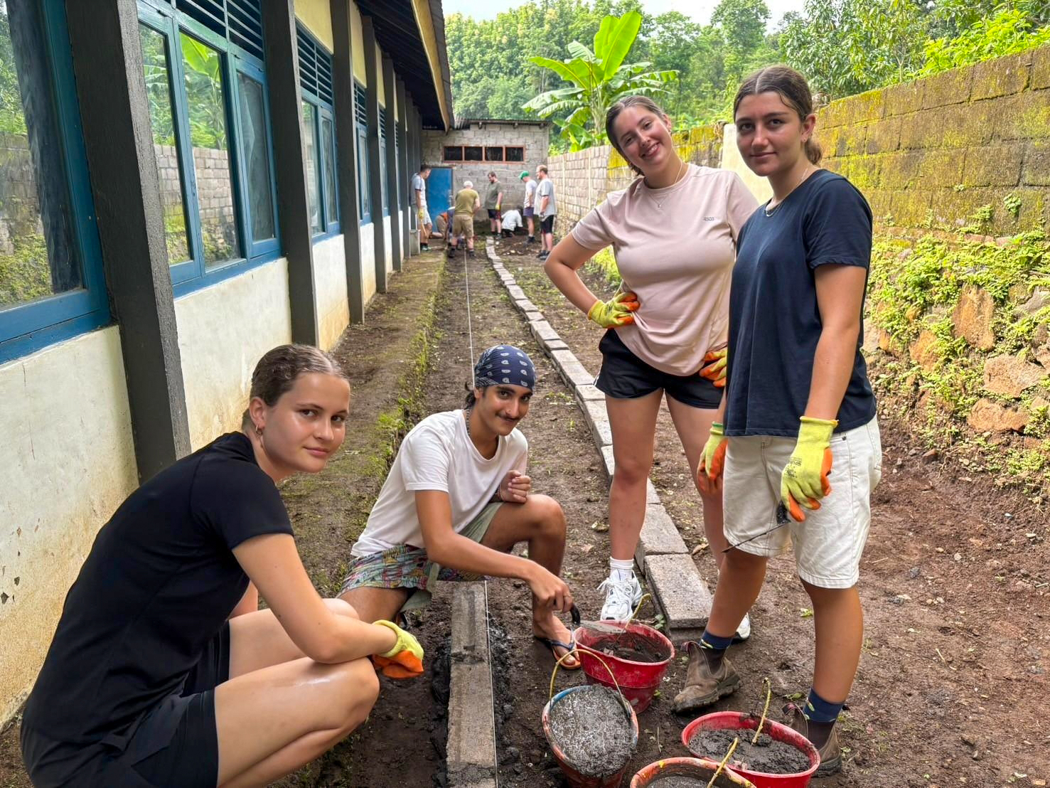 Four volunteers in work gloves smile beside the new concrete edging of a school path.