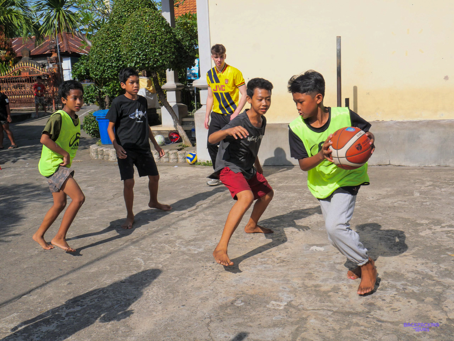 Boys in green bibs play basketball on a concrete court while a volunteer coach looks on.