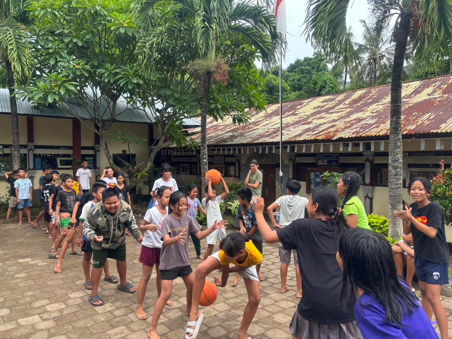 A crowd of children with basketballs gathers under the palm trees outside the school.