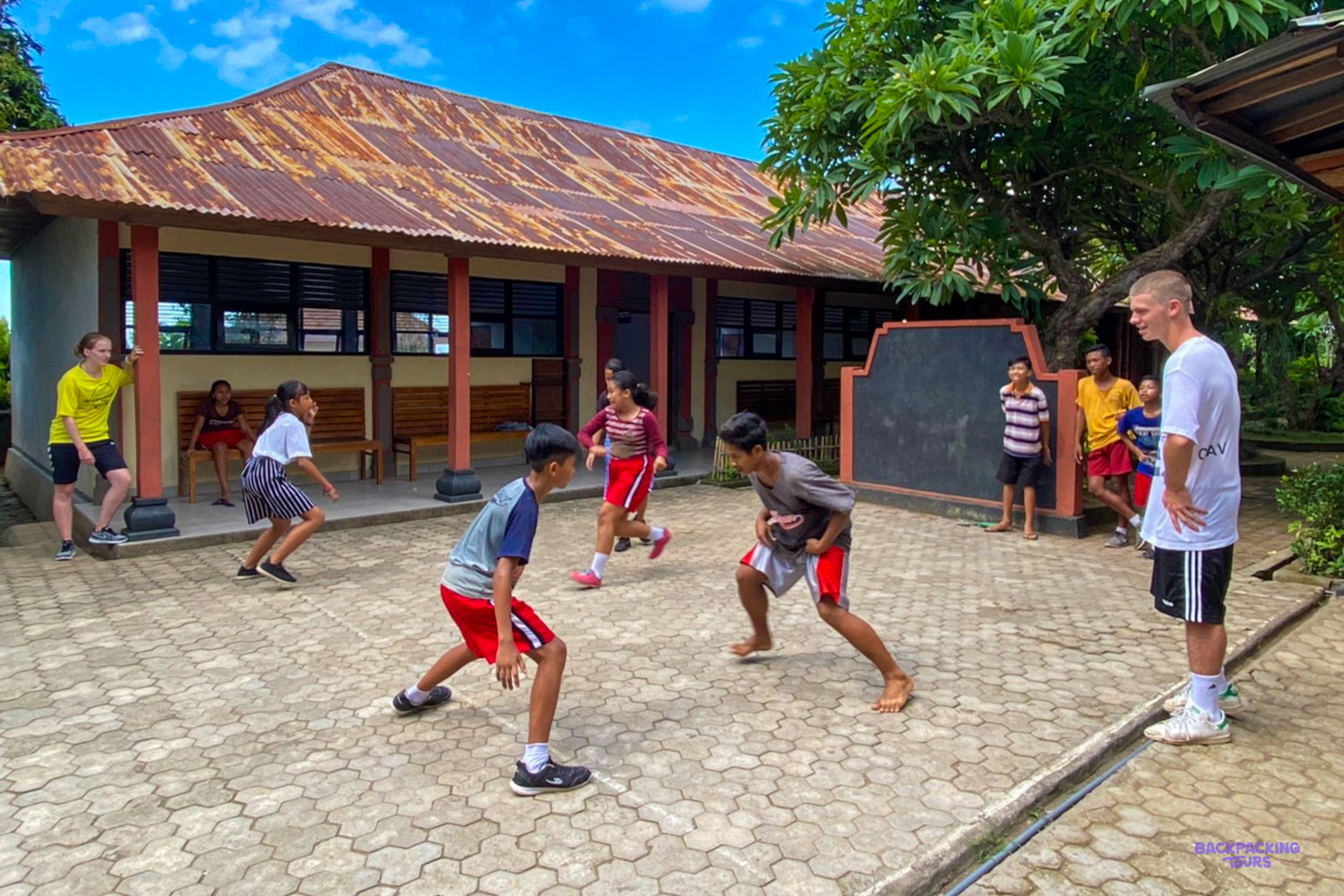 A volunteer coach watches children play ball in a paved schoolyard in front of the classrooms.