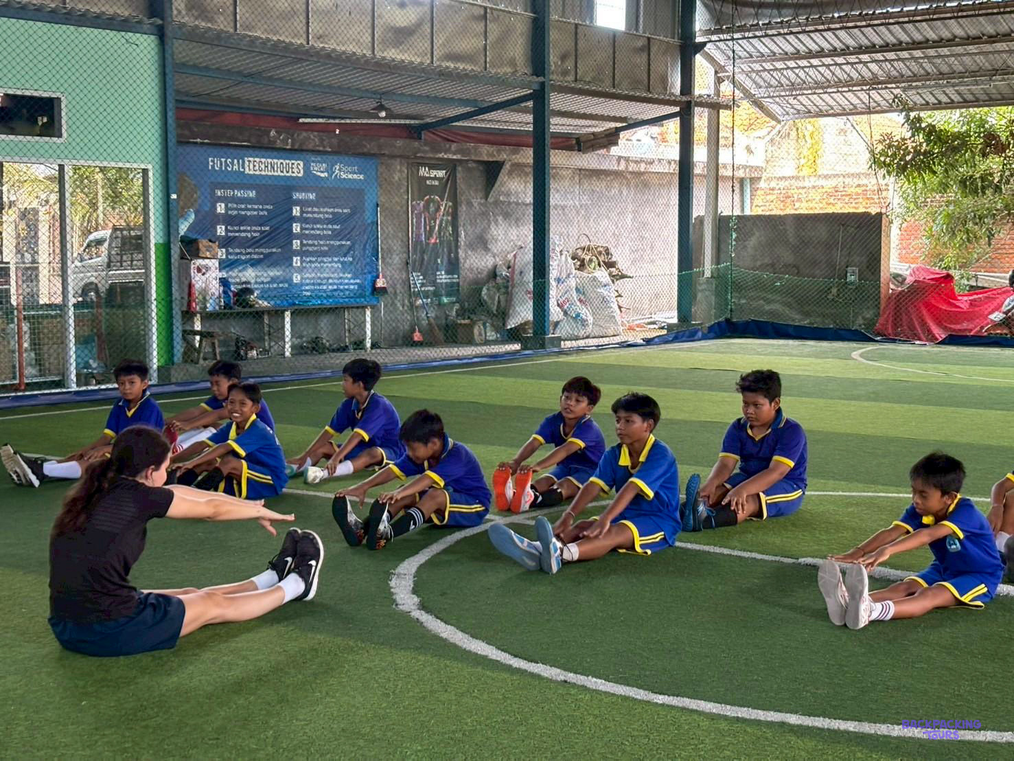 A volunteer leads boys in blue kit through stretches on an indoor futsal pitch.