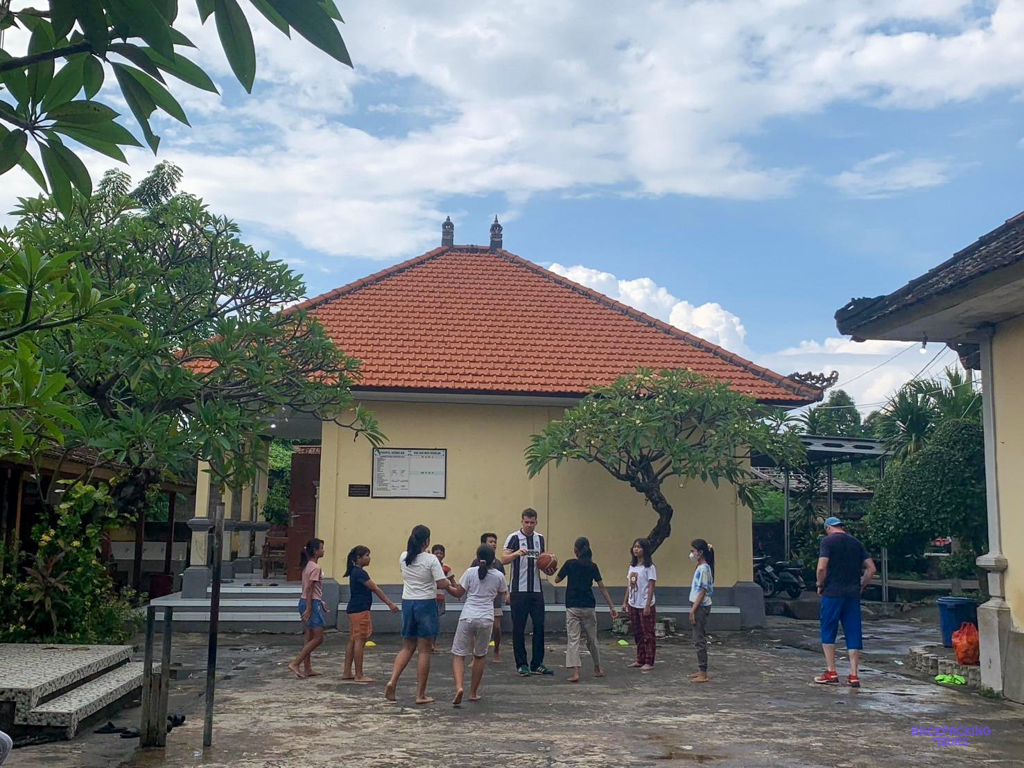 Children and volunteers play a game in the courtyard of a tiled-roof school building.