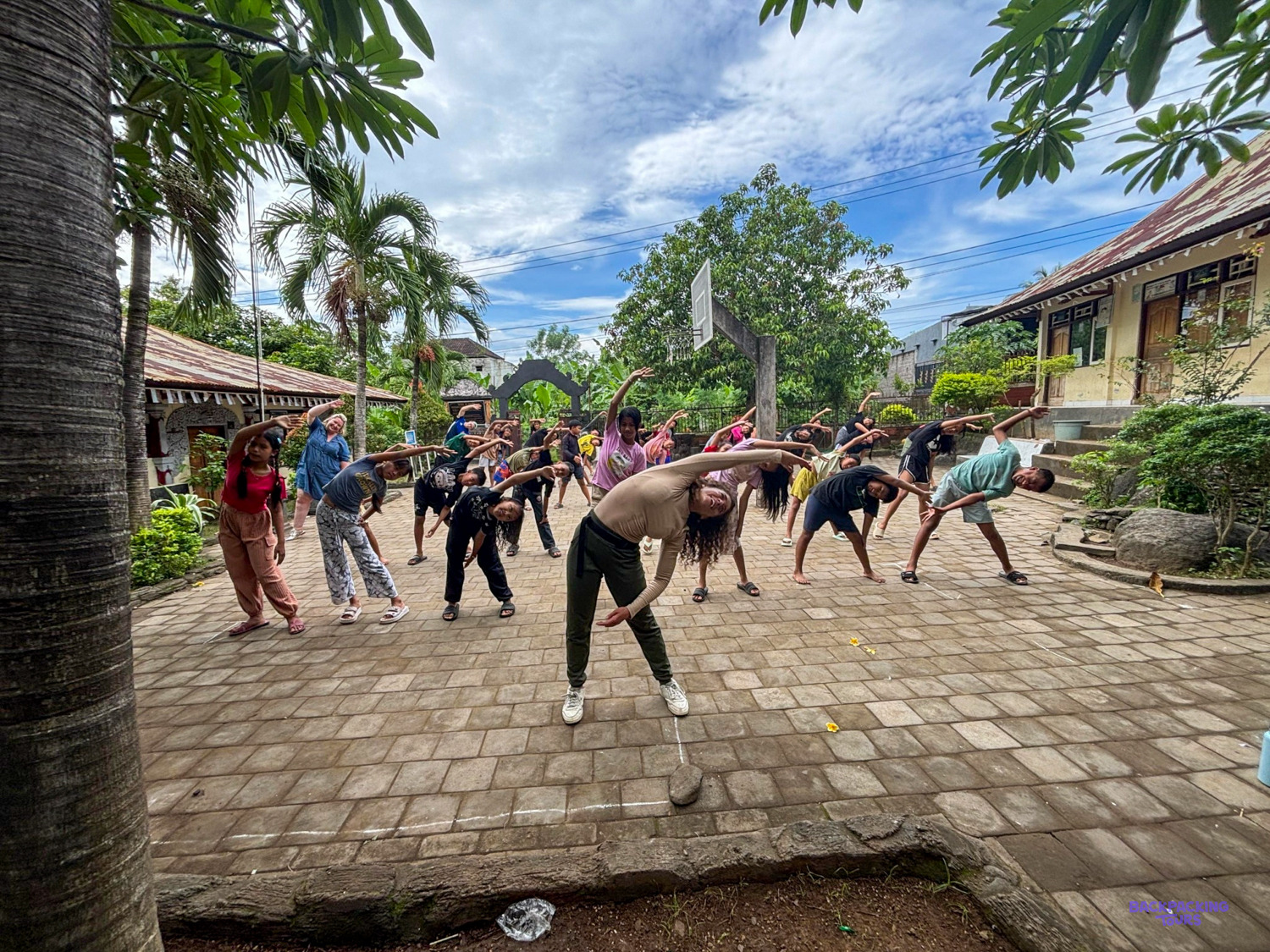 A volunteer leads a big group of children in side stretches in a palm-shaded courtyard.