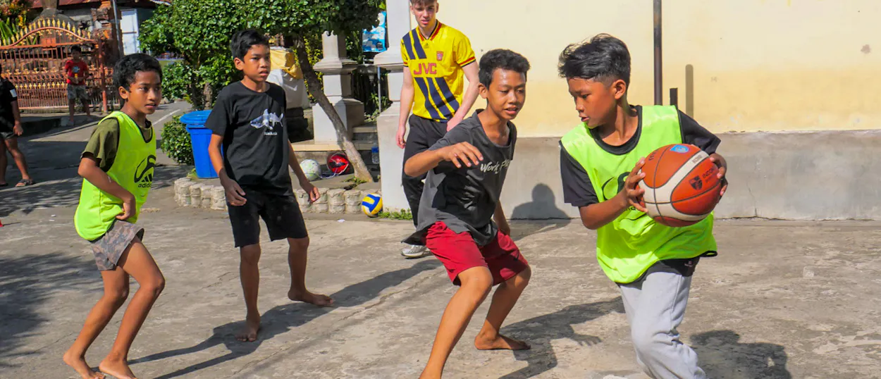Boys in green bibs play basketball on a concrete court while a volunteer coach looks on.