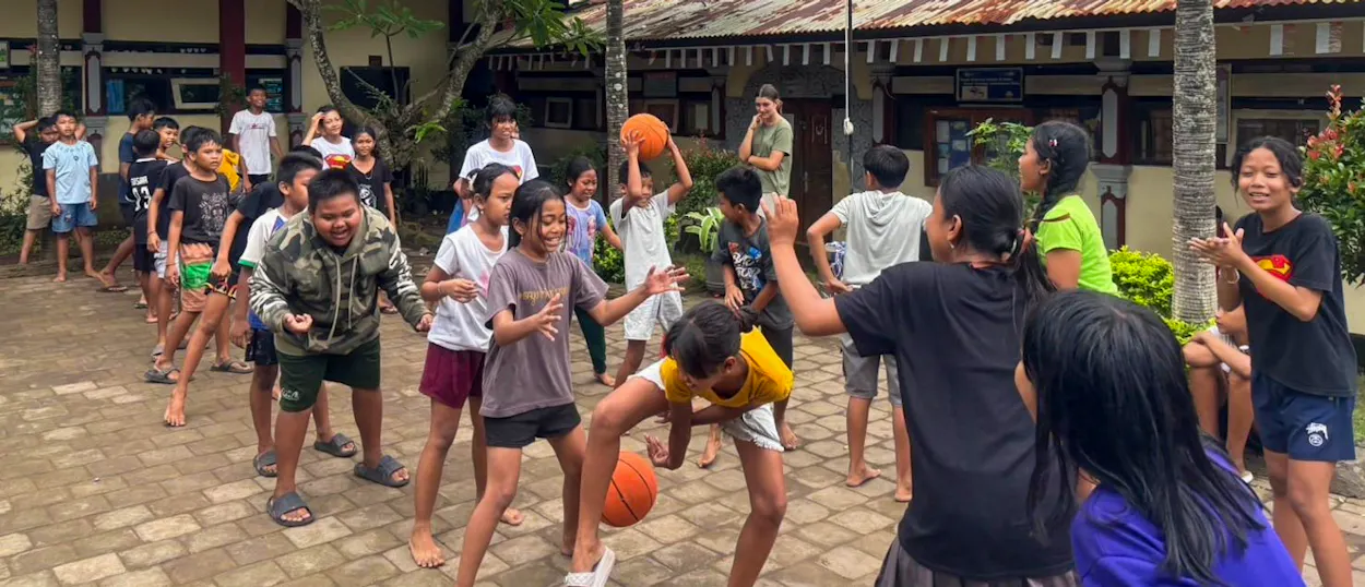A crowd of children with basketballs gathers under the palm trees outside the school.