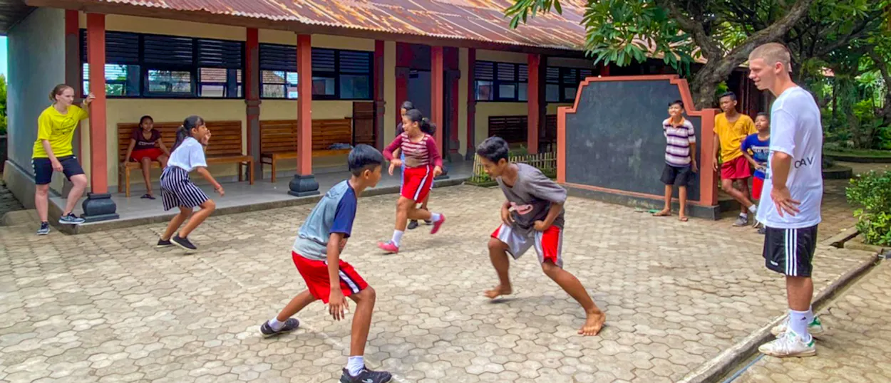 A volunteer coach watches children play ball in a paved schoolyard in front of the classrooms.