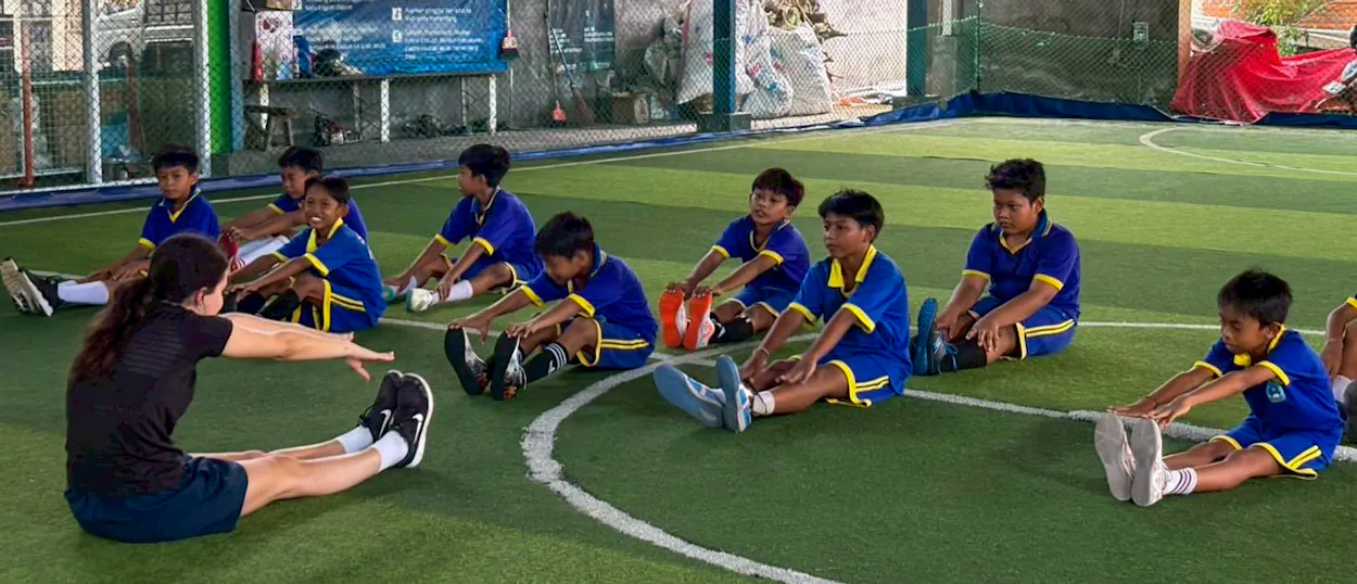 A volunteer leads boys in blue kit through stretches on an indoor futsal pitch.