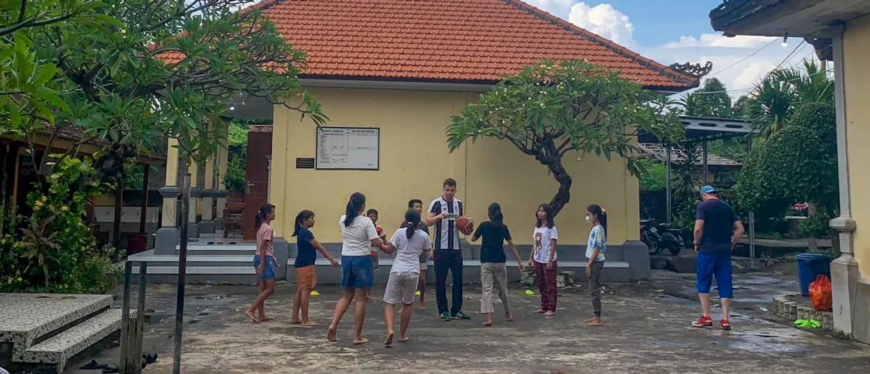 Children and volunteers play a game in the courtyard of a tiled-roof school building.