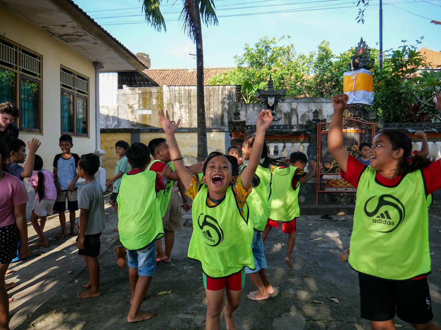 Children in green training bibs cheer with their arms in the air in a school courtyard.