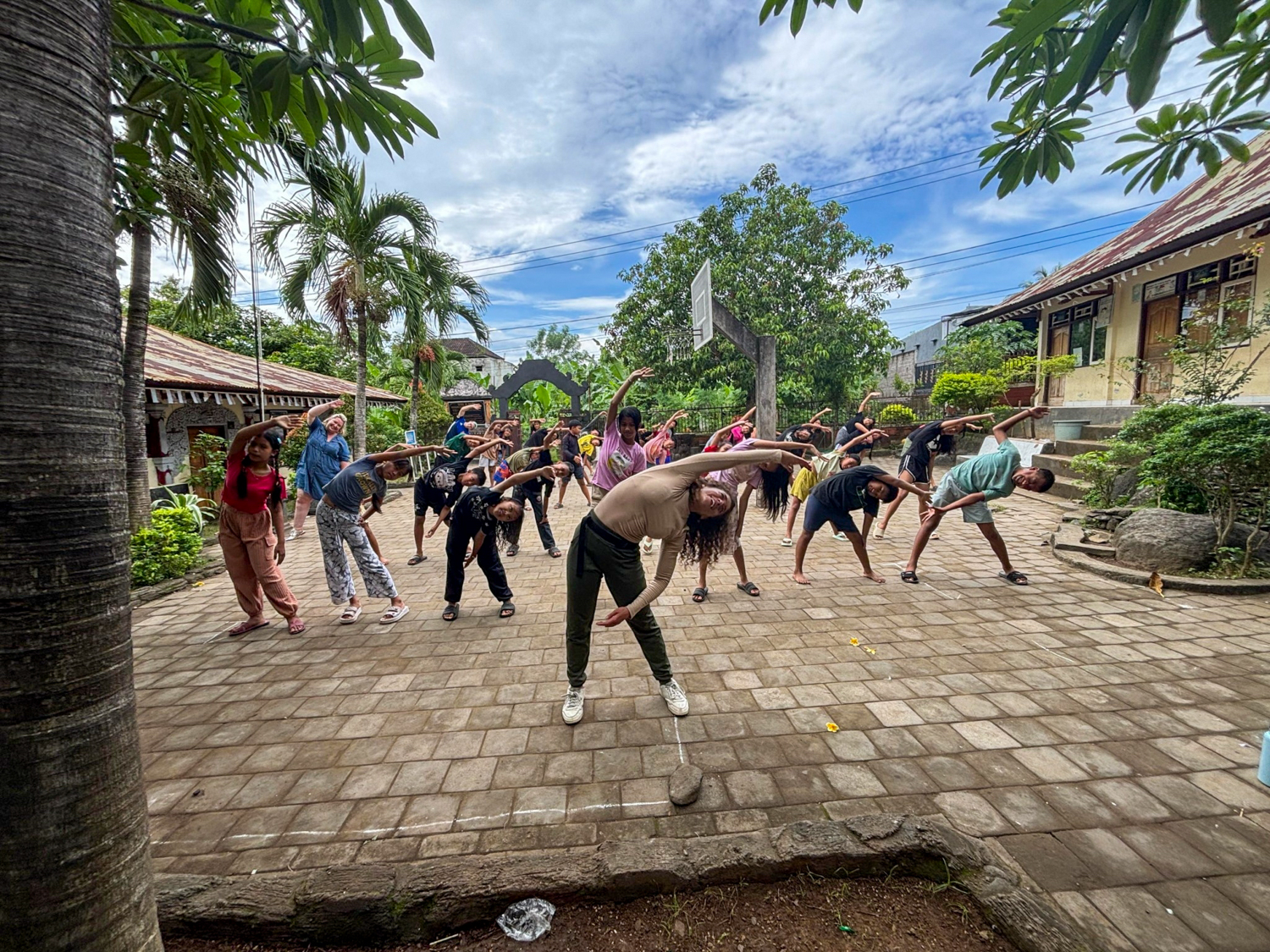 A volunteer leads a big group of children in side stretches in a palm-shaded courtyard.