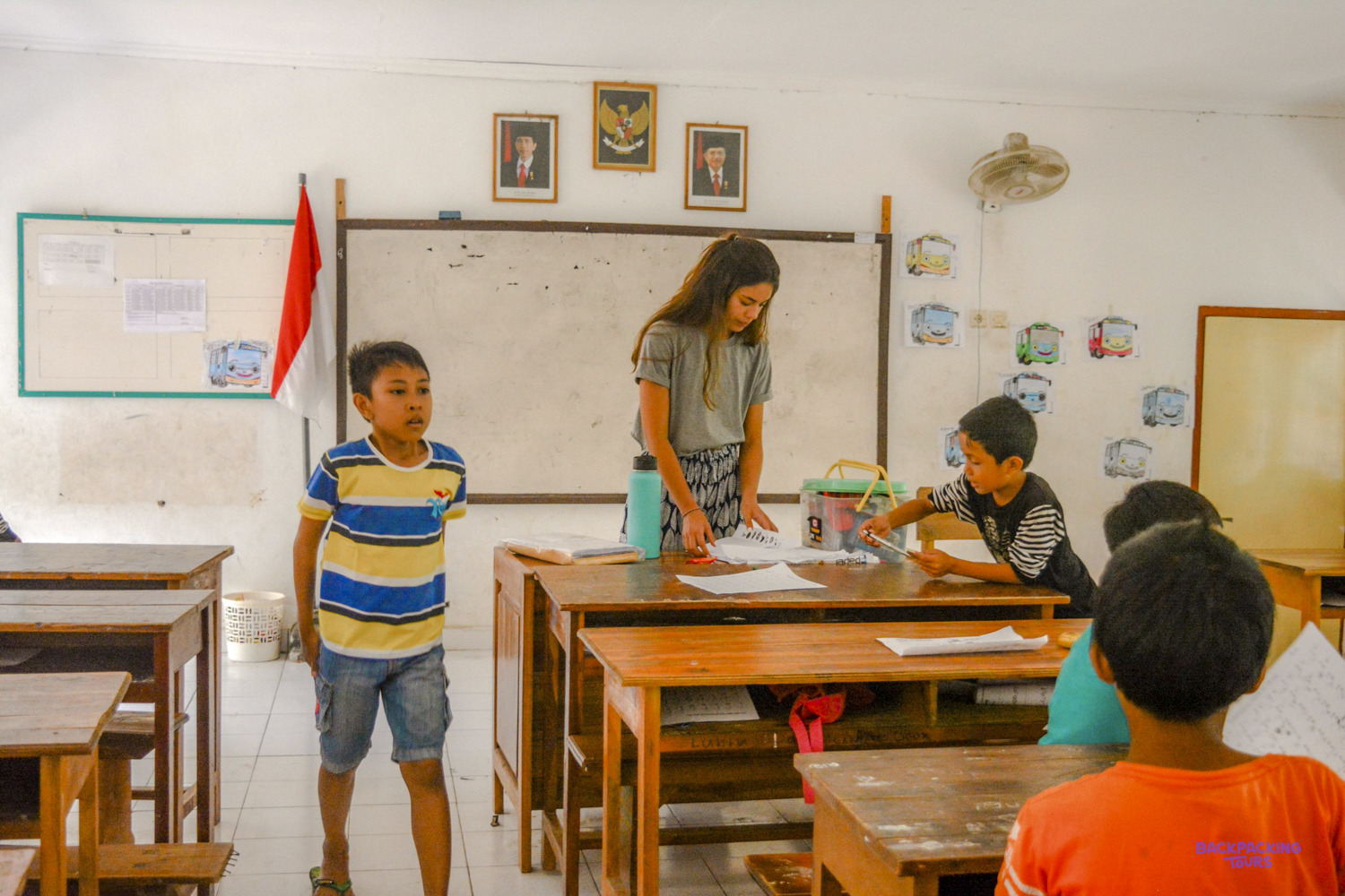 A volunteer sorts worksheets at a classroom desk while a boy stands up to answer.