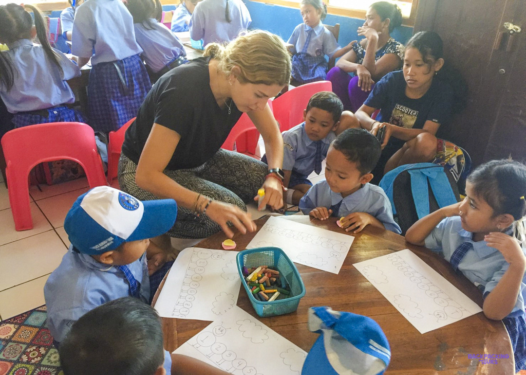 A volunteer kneels at a low table, helping young pupils with their colouring worksheets.