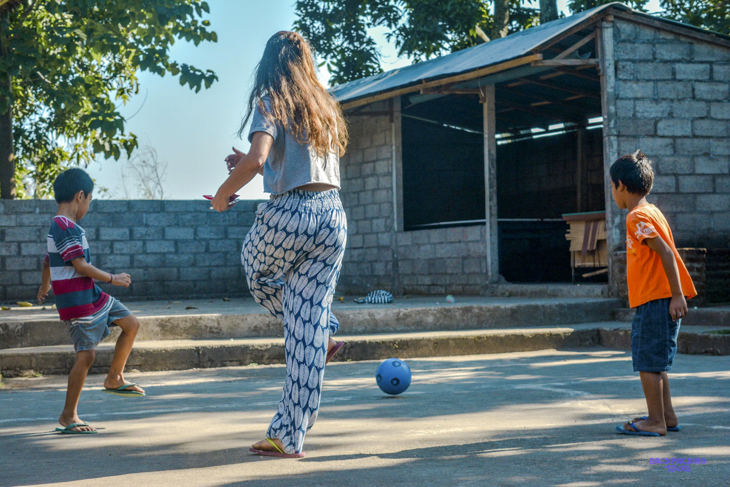 A volunteer kicks a football around the sunny schoolyard with two young boys.