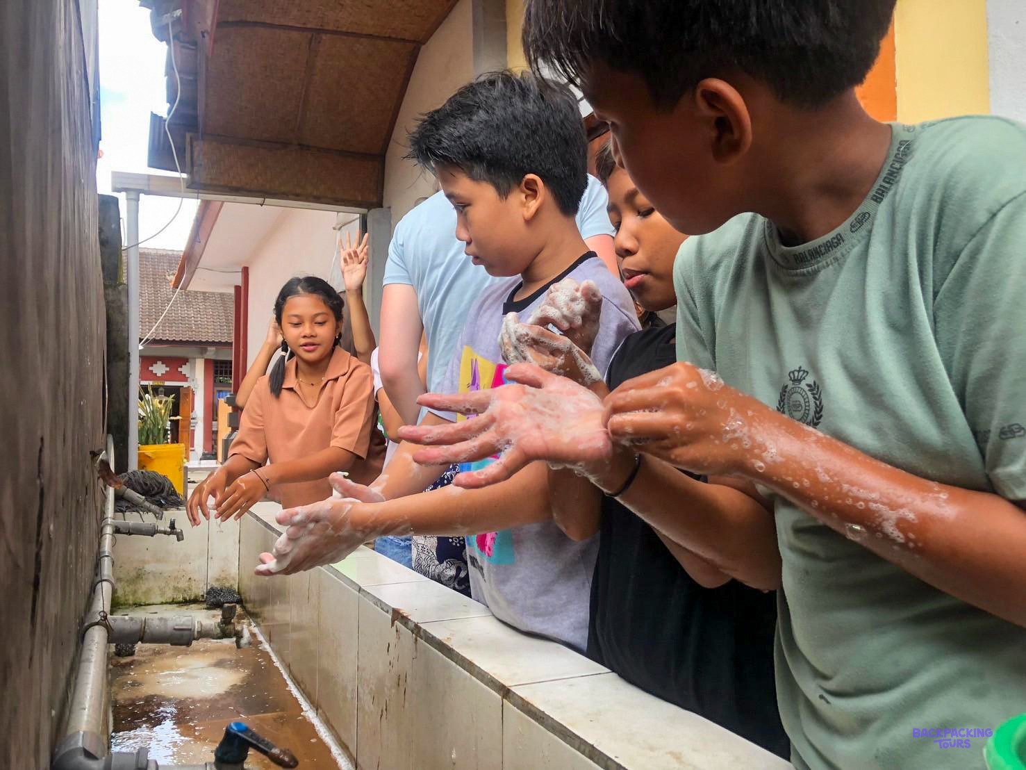 Children rinse their soapy hands at an outdoor school sink before class.