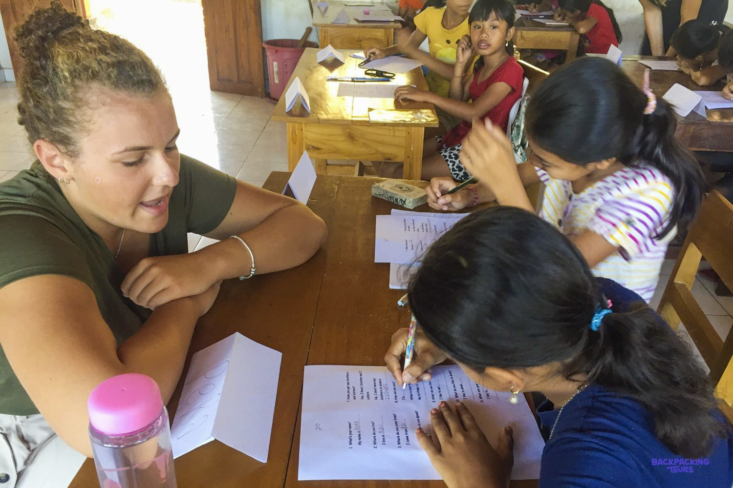 A volunteer leans in to help two girls write out their English words at a classroom desk.