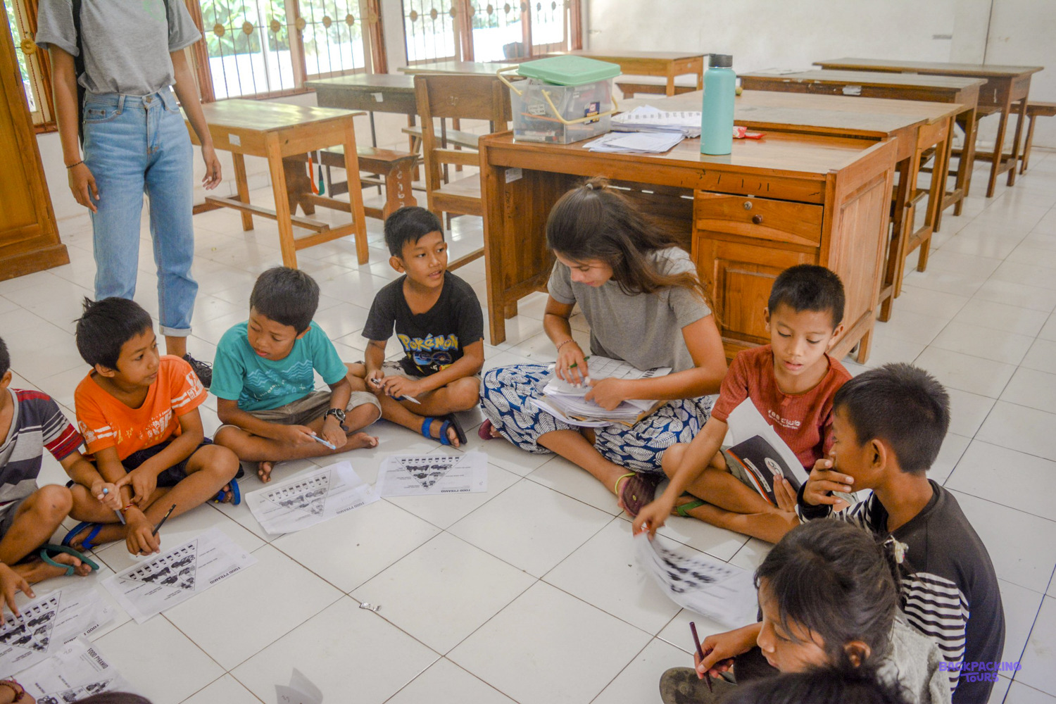 A volunteer sits on the classroom floor in a circle of children working on their worksheets.