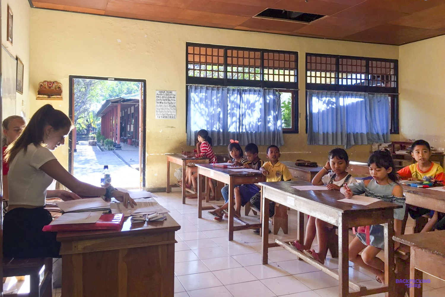 A volunteer prepares her lesson at the teacher's desk while children wait at their desks.