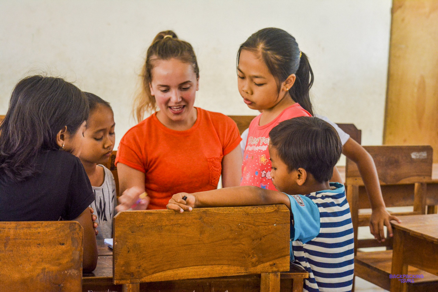 A smiling volunteer in an orange top works through a worksheet with a group of children.