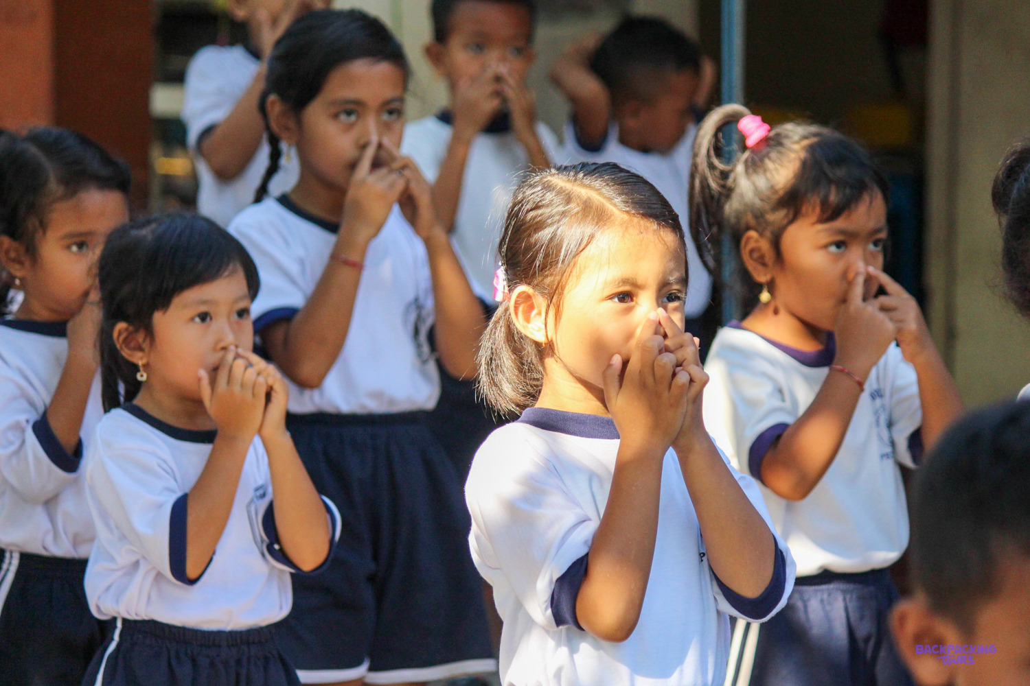 Young schoolchildren in white uniforms giggle behind their hands during a classroom game.
