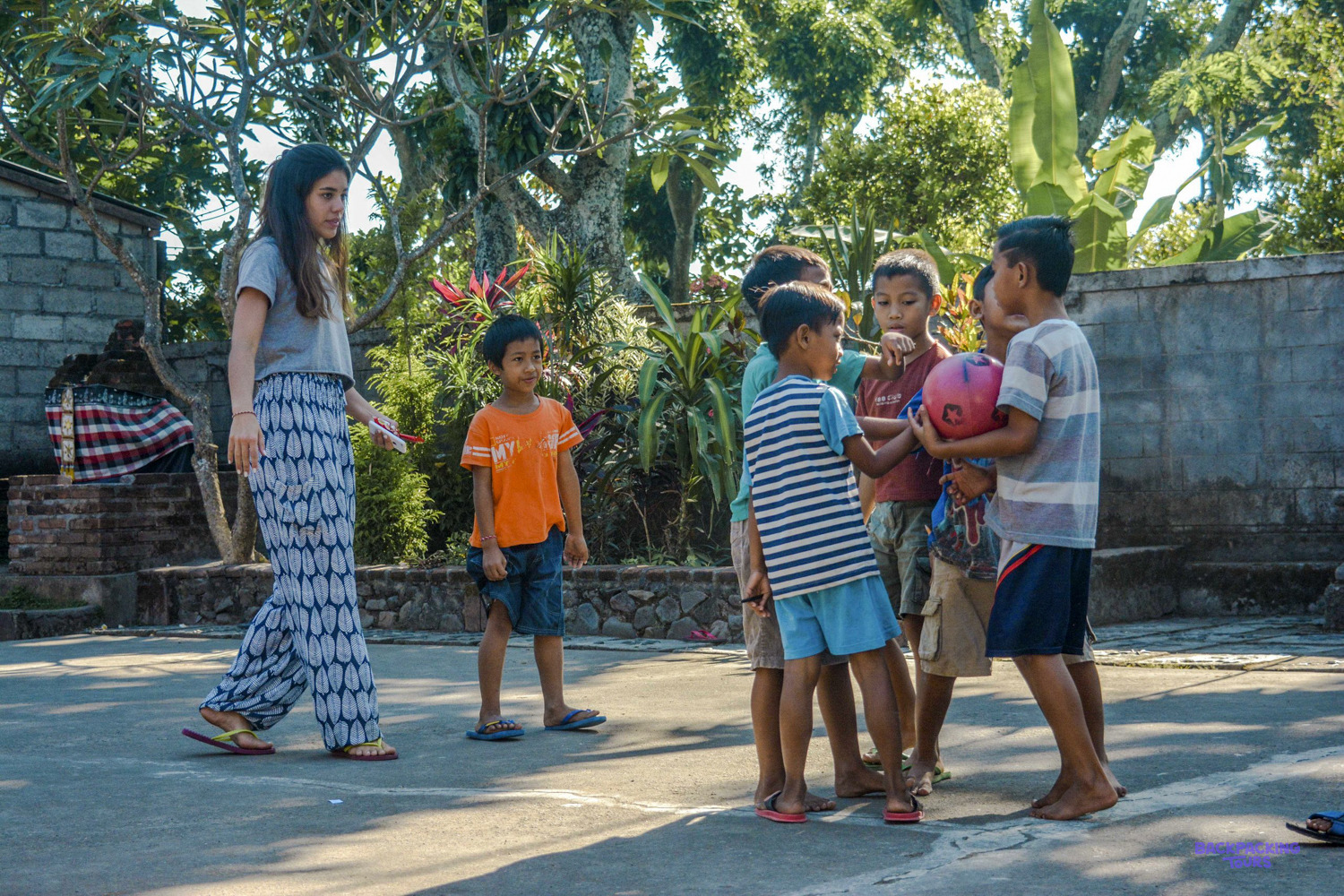 A volunteer joins a group of boys holding a red ball in a sunny schoolyard.