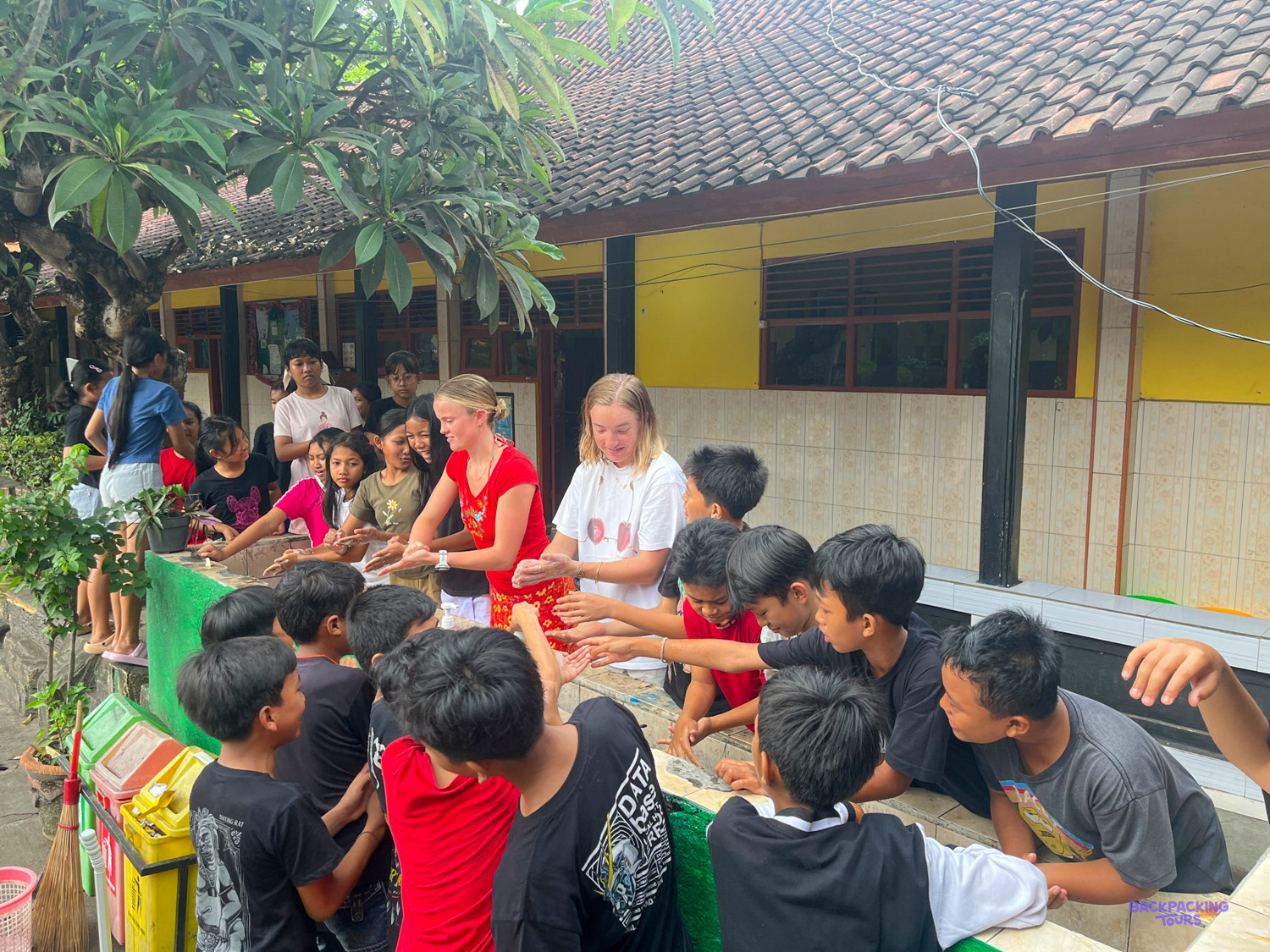 Two volunteers lead a crowd of schoolchildren around an outdoor table in a school courtyard.