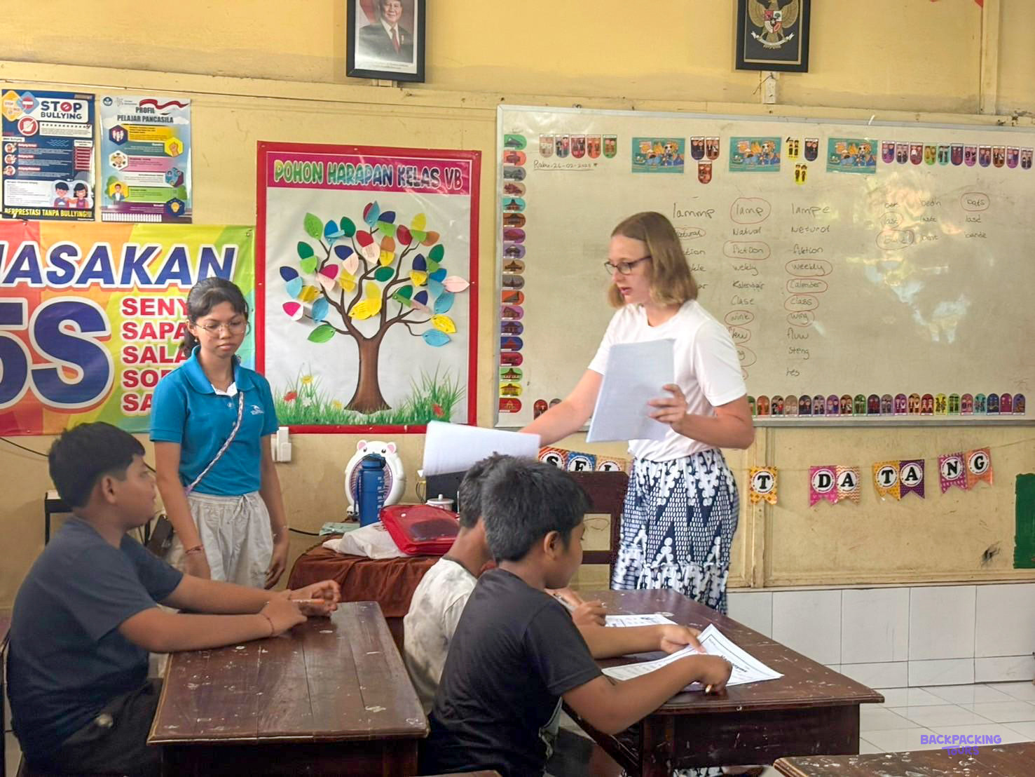 A volunteer reads out a worksheet beside a local teacher in a colourful primary classroom.
