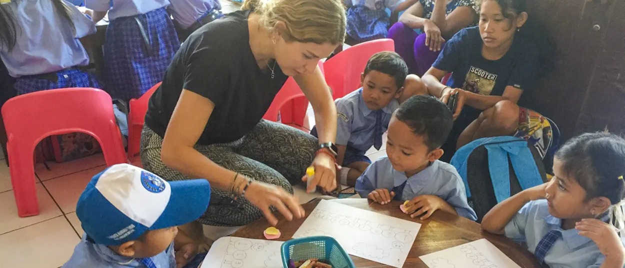 A volunteer kneels at a low table, helping young pupils with their colouring worksheets.
