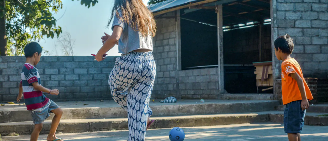 A volunteer kicks a football around the sunny schoolyard with two young boys.