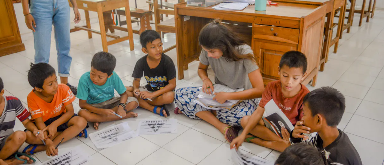 A volunteer sits on the classroom floor in a circle of children working on their worksheets.