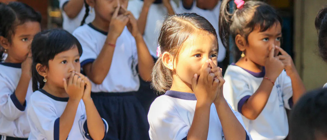 Young schoolchildren in white uniforms giggle behind their hands during a classroom game.
