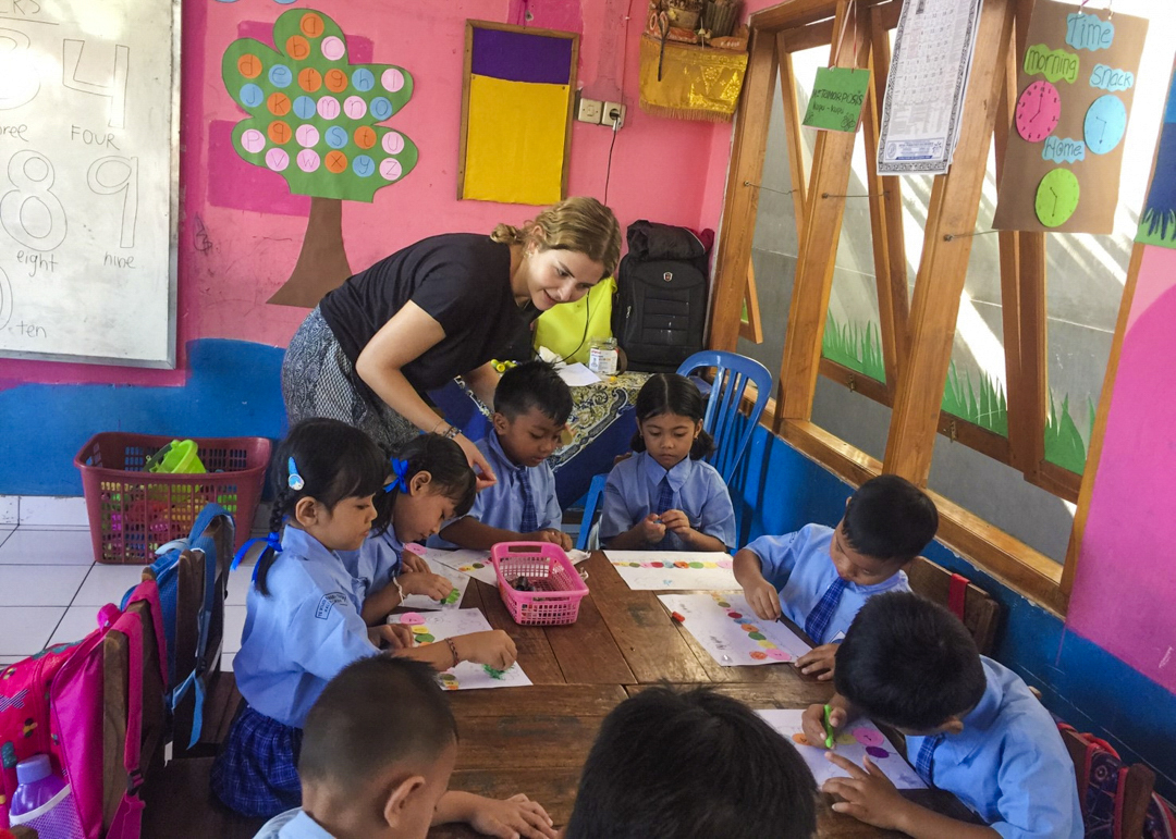 A volunteer leans over a table of kindergarten children colouring in a bright pink classroom.