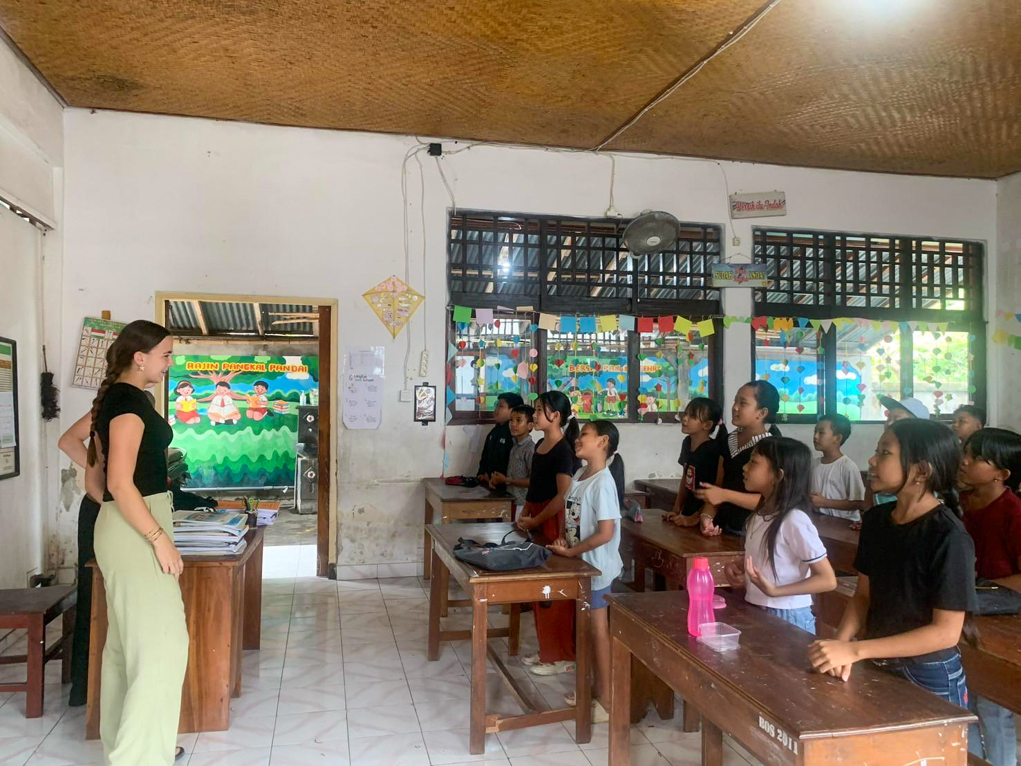 A volunteer stands at the front of a bright classroom, teaching a full class of children.