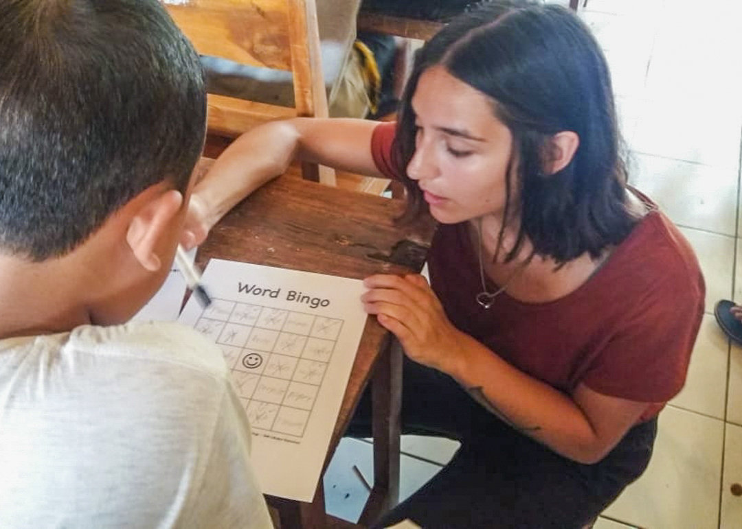 A volunteer kneels beside a boy's desk to help him through a game of word bingo.