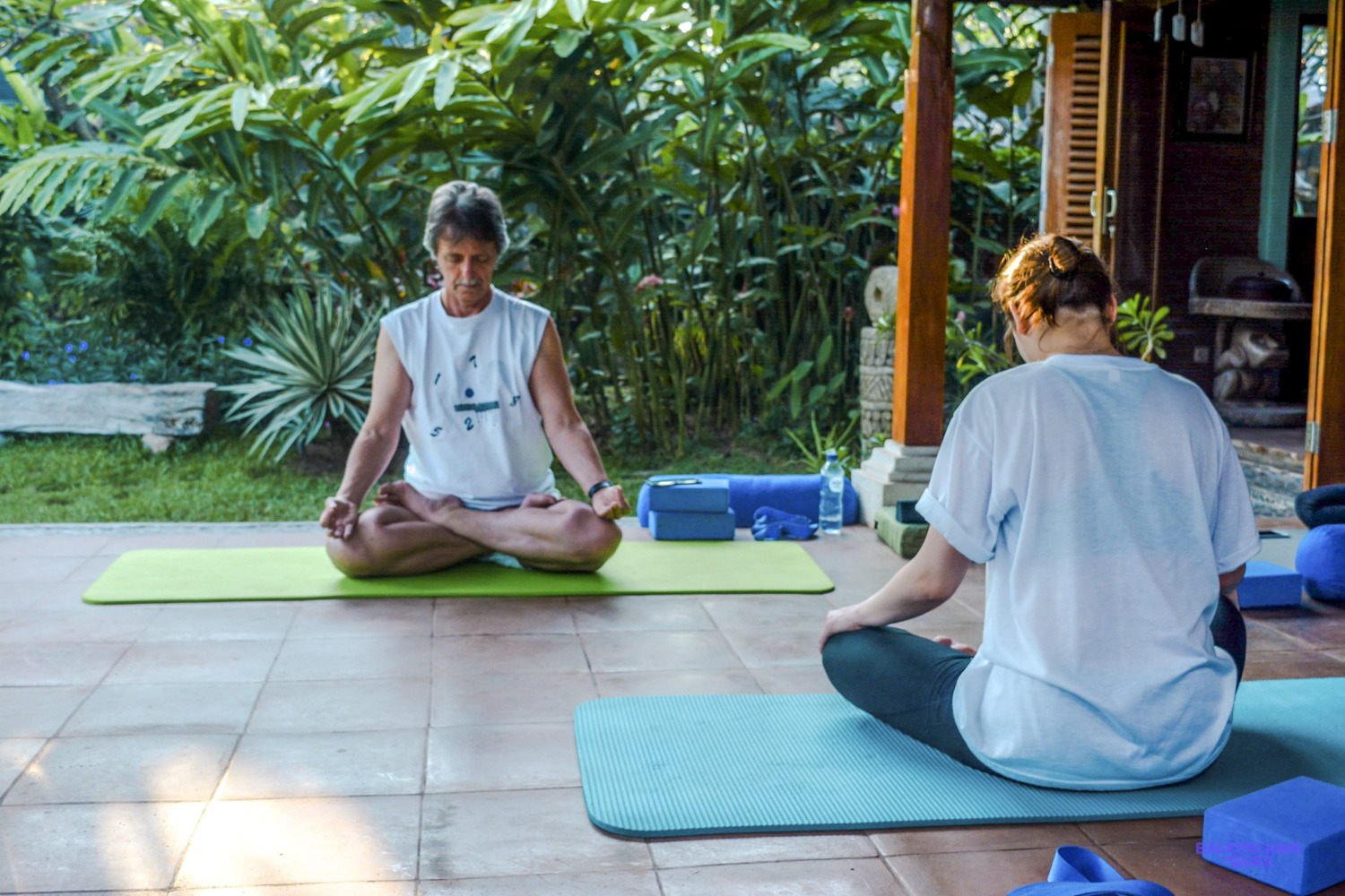 Two people sit cross-legged on yoga mats on a tiled terrace, meditating beside a leafy garden.
