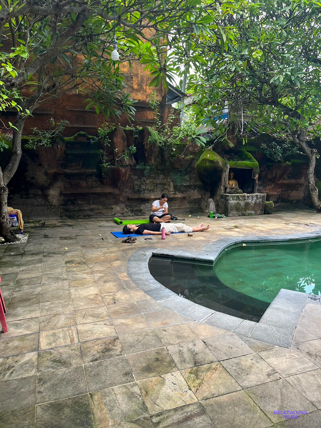 Two people on yoga mats by a curved pool in a shady garden of stone walls and trees.