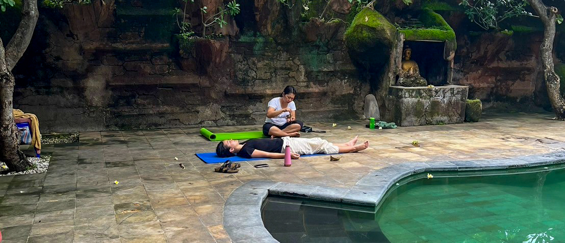 Two people on yoga mats by a curved pool in a shady garden of stone walls and trees.