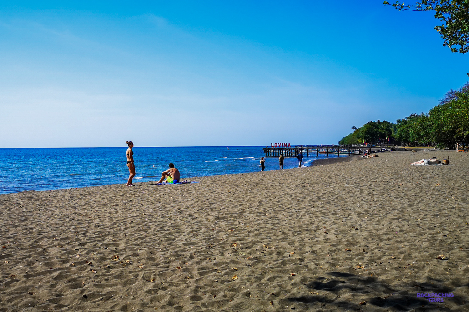 Swimmers on a wide black-sand beach under a clear blue sky, with a jetty in the distance.