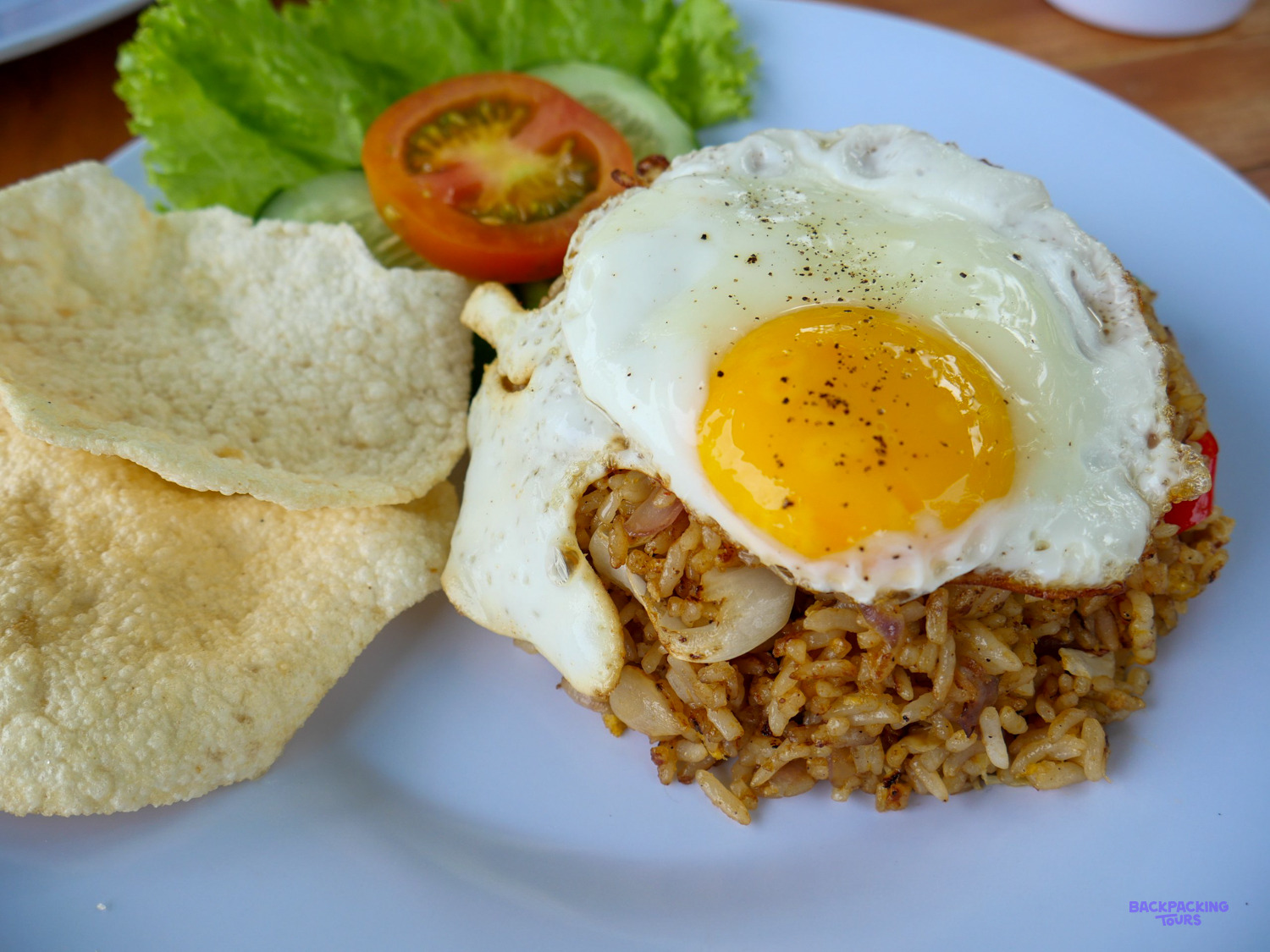 A plate of nasi goreng topped with a fried egg, prawn crackers, tomato and cucumber.