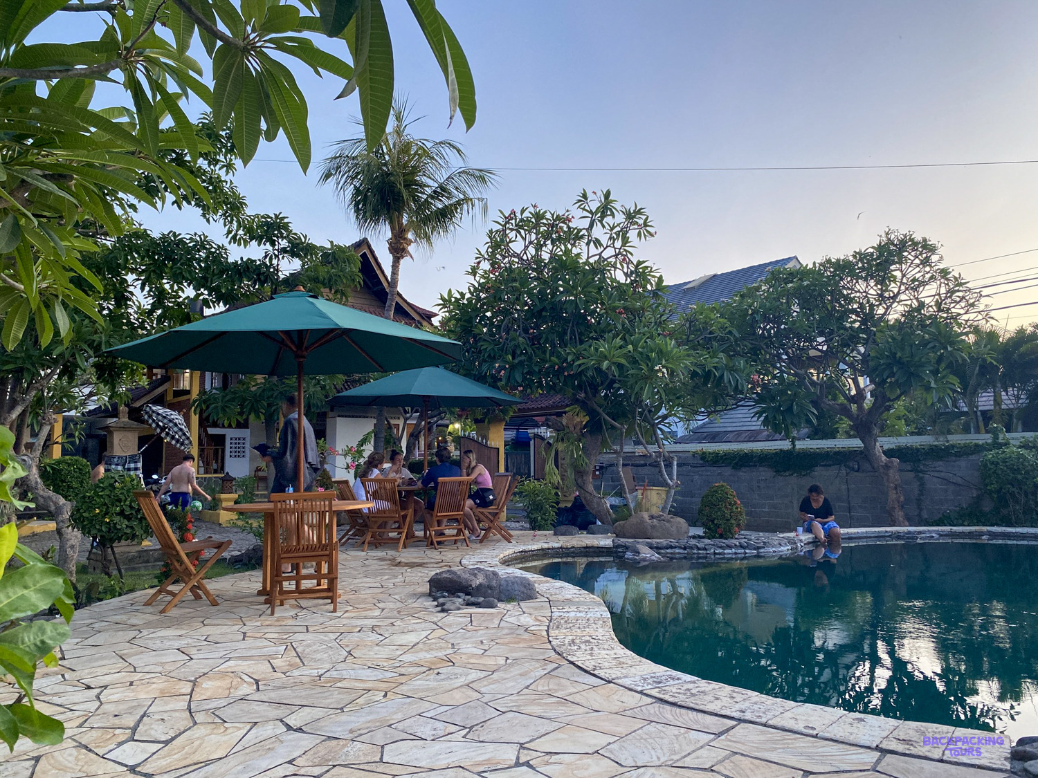 Garden tables under green parasols beside the pool, with frangipani trees and palms behind.