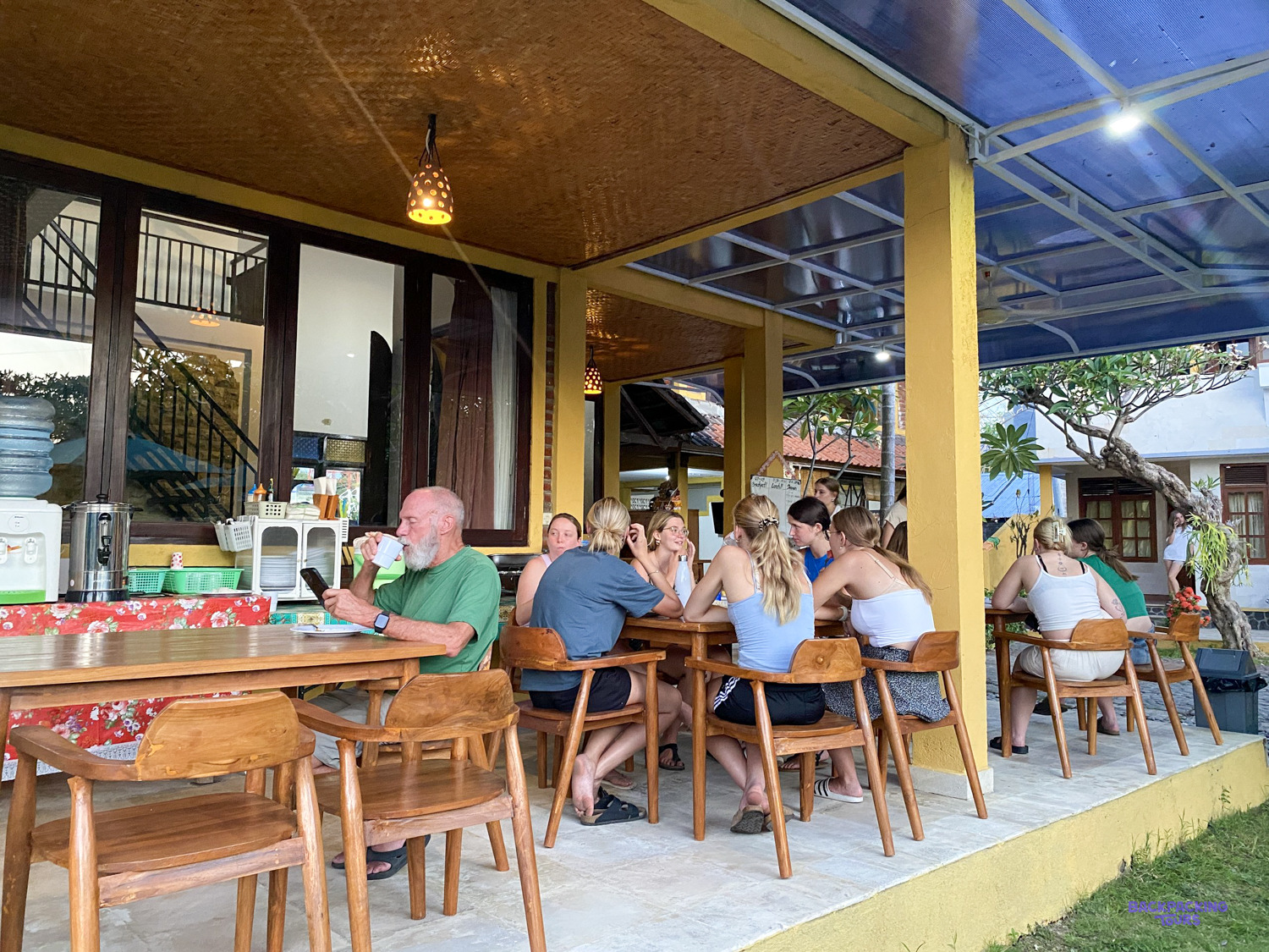 Volunteers chat over a meal at long wooden tables on the house's open terrace.