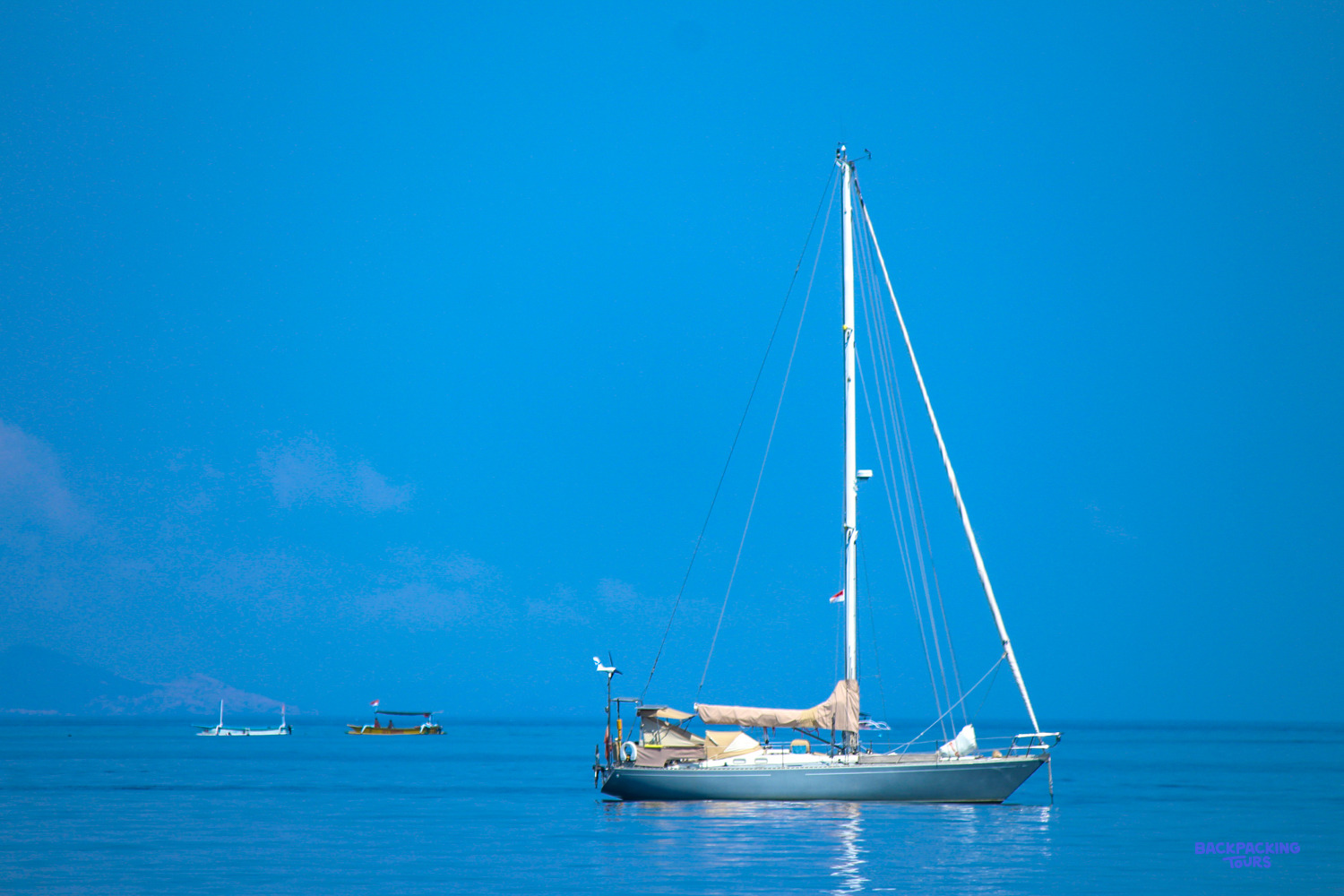 A white sailing yacht rides at anchor on a calm, bright blue sea off Lovina.