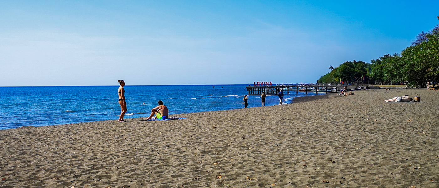 Swimmers on a wide black-sand beach under a clear blue sky, with a jetty in the distance.