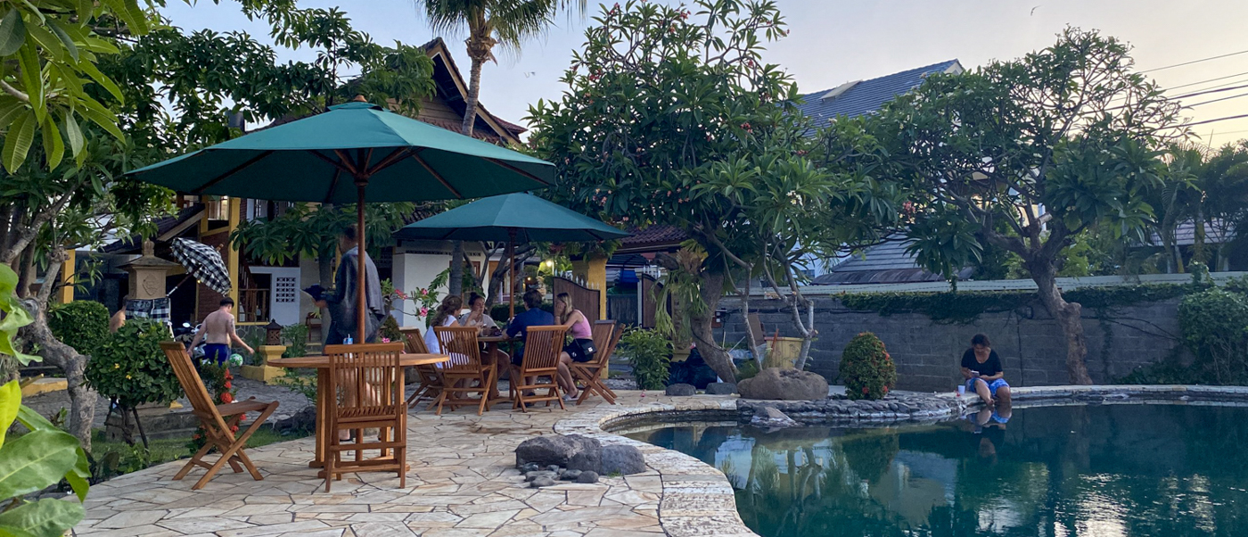 Garden tables under green parasols beside the pool, with frangipani trees and palms behind.