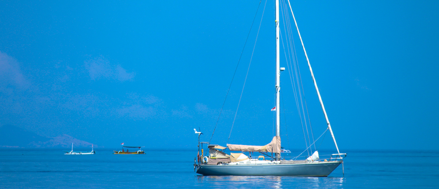 A white sailing yacht rides at anchor on a calm, bright blue sea off Lovina.